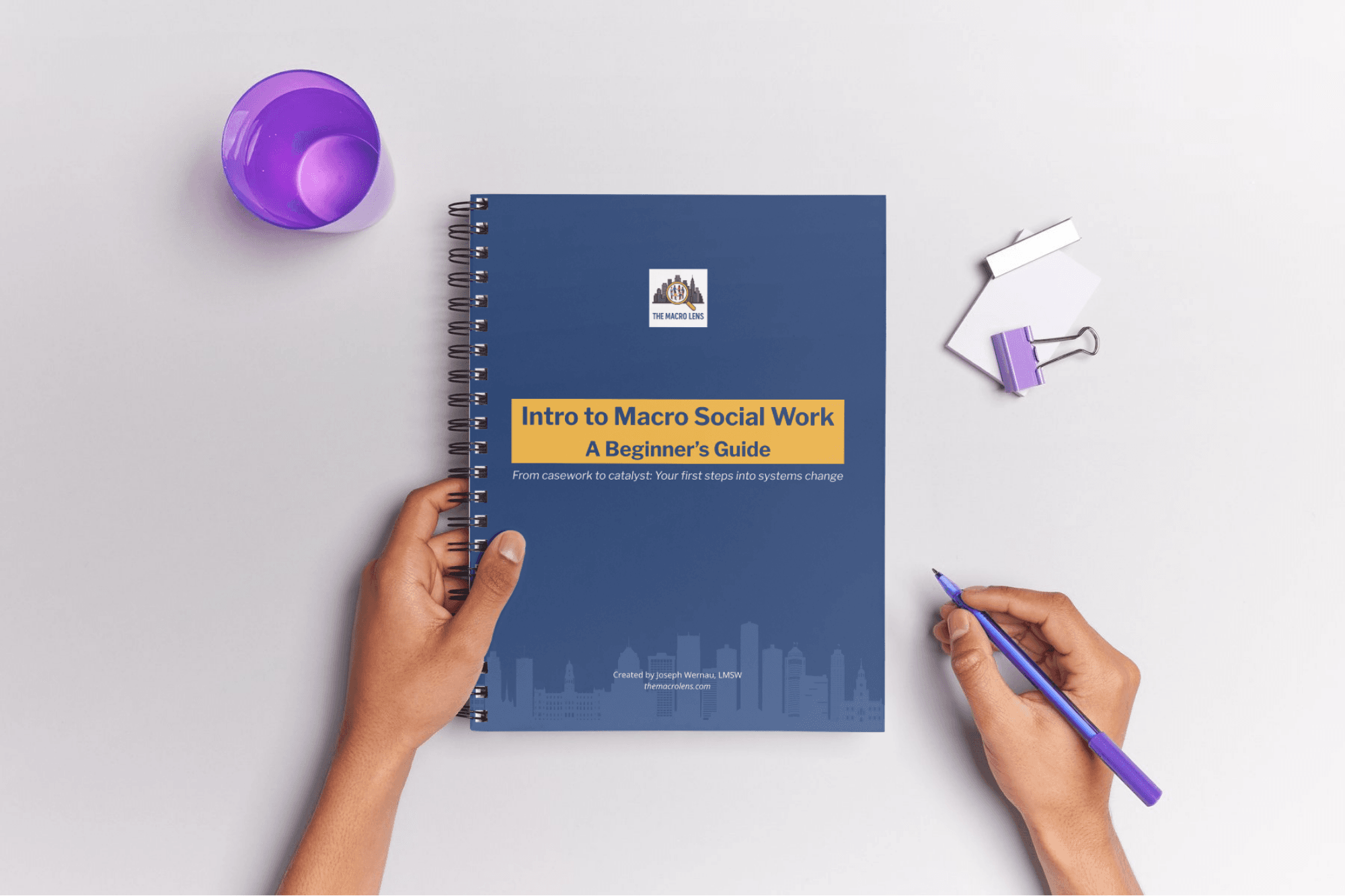 Hands holding pen beside spiral-bound workbook titled ‘Intro to Macro Social Work: A Beginner’s Guide’ on white desk with office supplies. Represents the free guide received when you subscribe to the macro social work newsletter.
